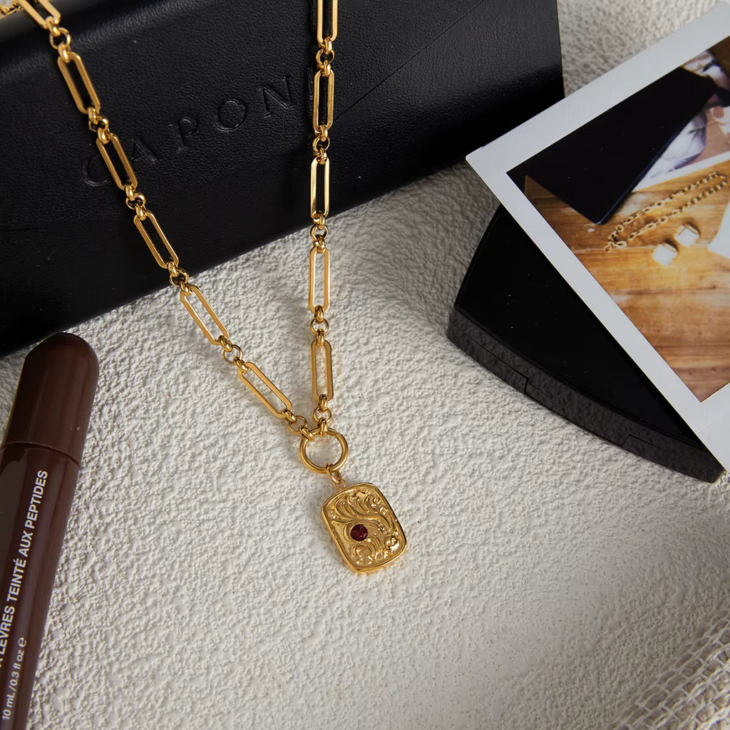 Gold coin necklace with red rhinestone detail as flatlay on white textured surface. 