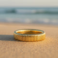 Gold ring with ribbed texture sitting on a sandy beach with blurred ocean in background