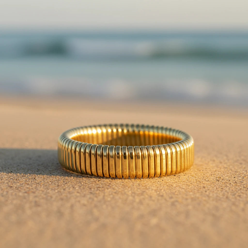 Gold ring with ribbed texture sitting on a sandy beach with blurred ocean in background