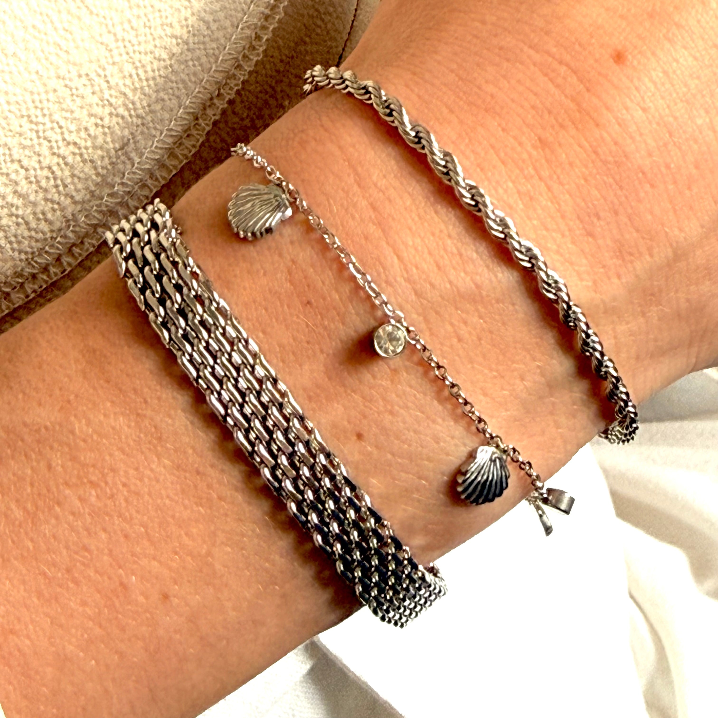 Close-up of a wrist wearing multiple silver bracelets with shell charms. rope chain bracelet silver, silver chain bracelet, textured bracelet