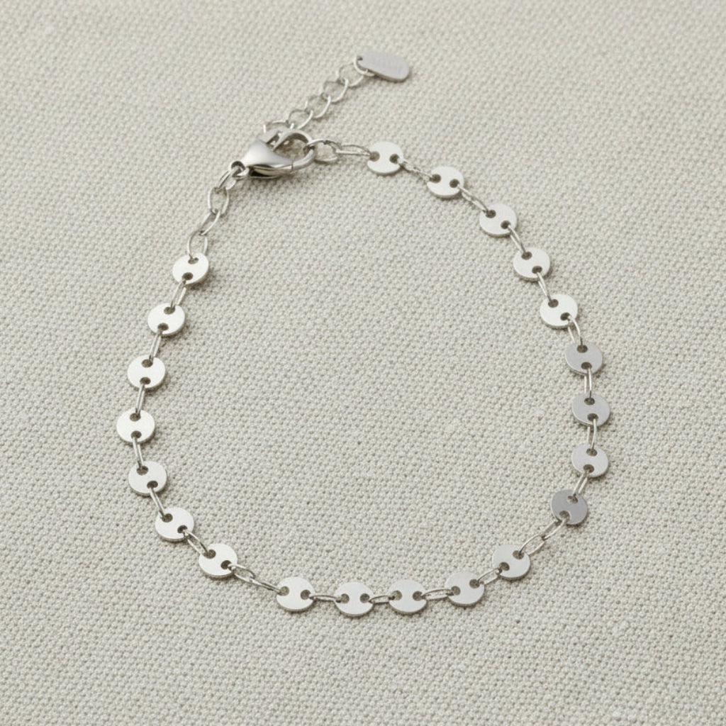 Silver disk bracelet on a white surface with a clear dish in the background