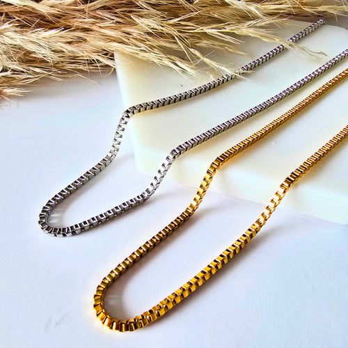 One gold and one silver box chain necklace on a white surface with pampas grass. 