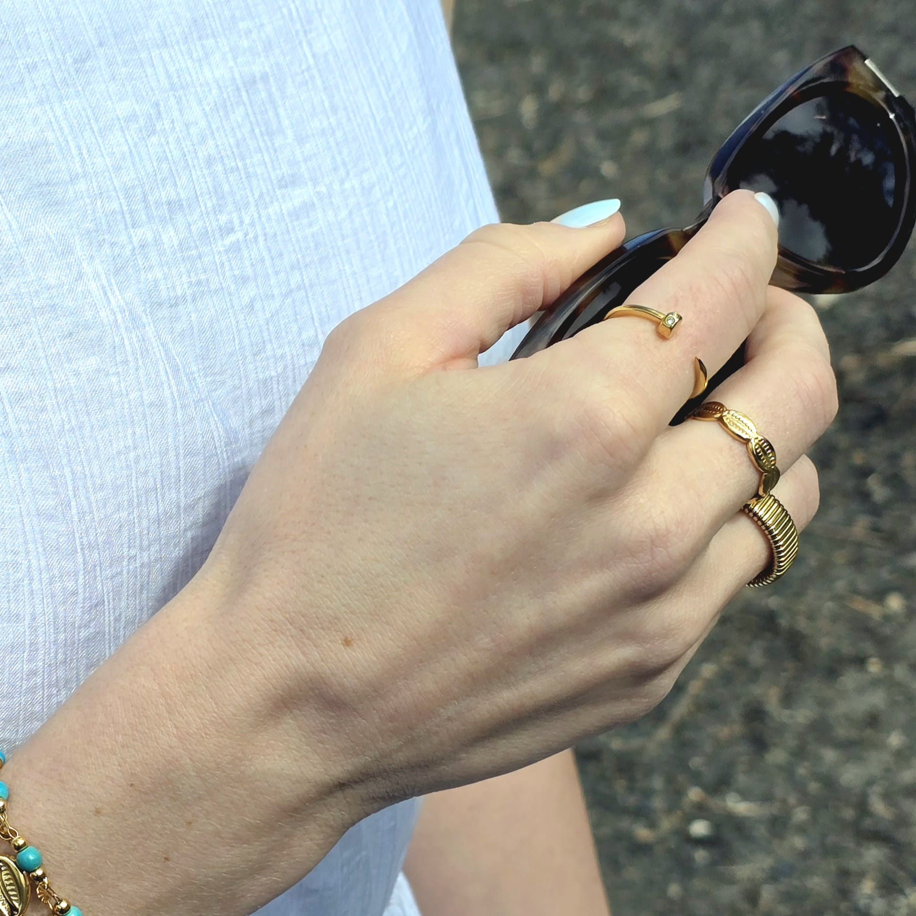 Hand holding sunglasses with gold rings and a bracelet on a blurred background